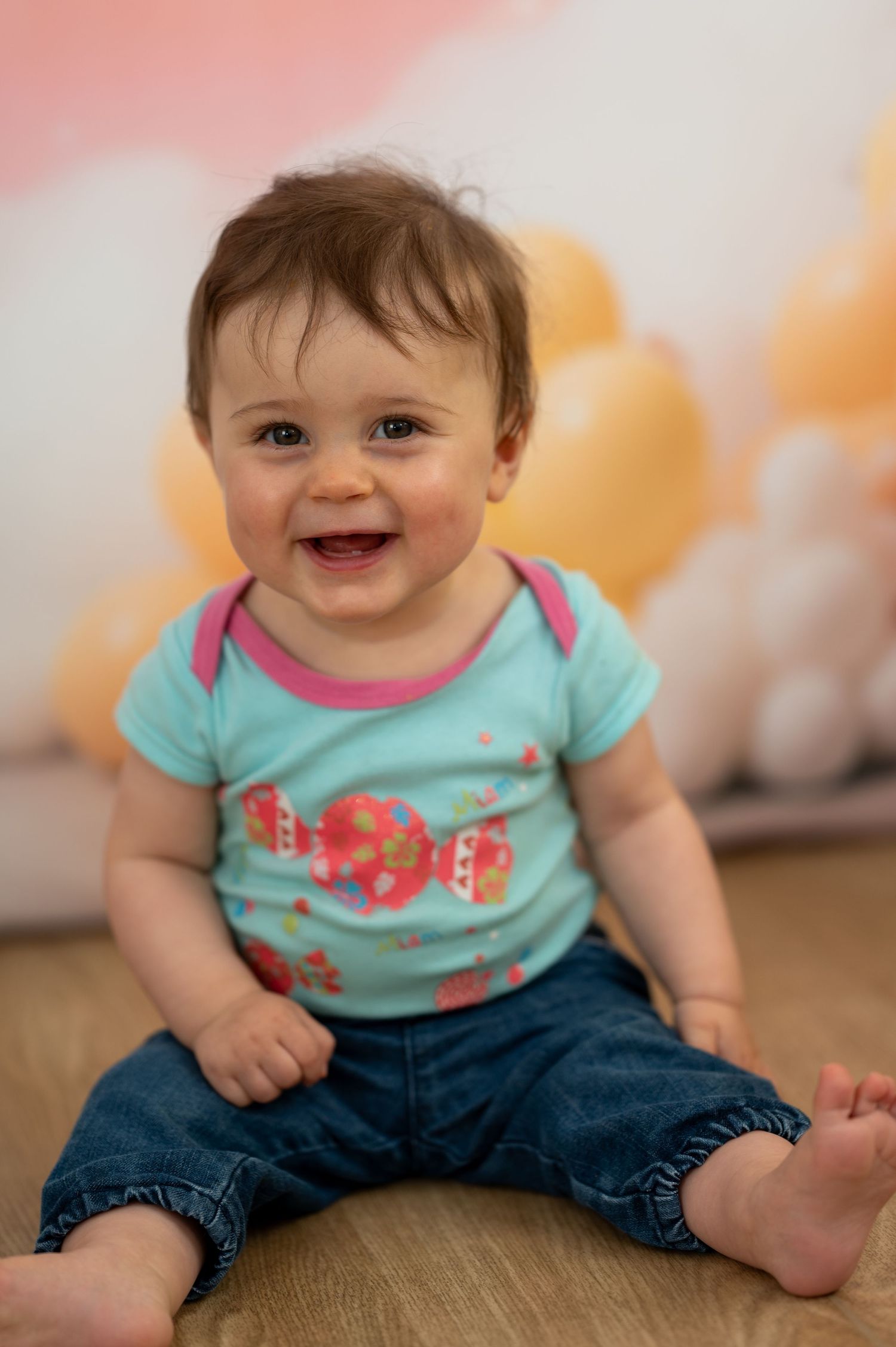 Smiling baby in teal floral onesie and jeans sitting on floor with yellow balloons in background.