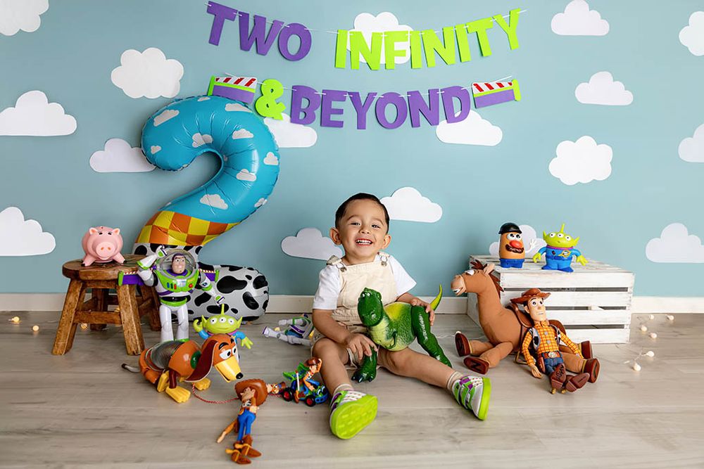 Toddler boy's Toy Story 2nd birthday photo with Buzz, Woody, and friends against a cloud backdrop.