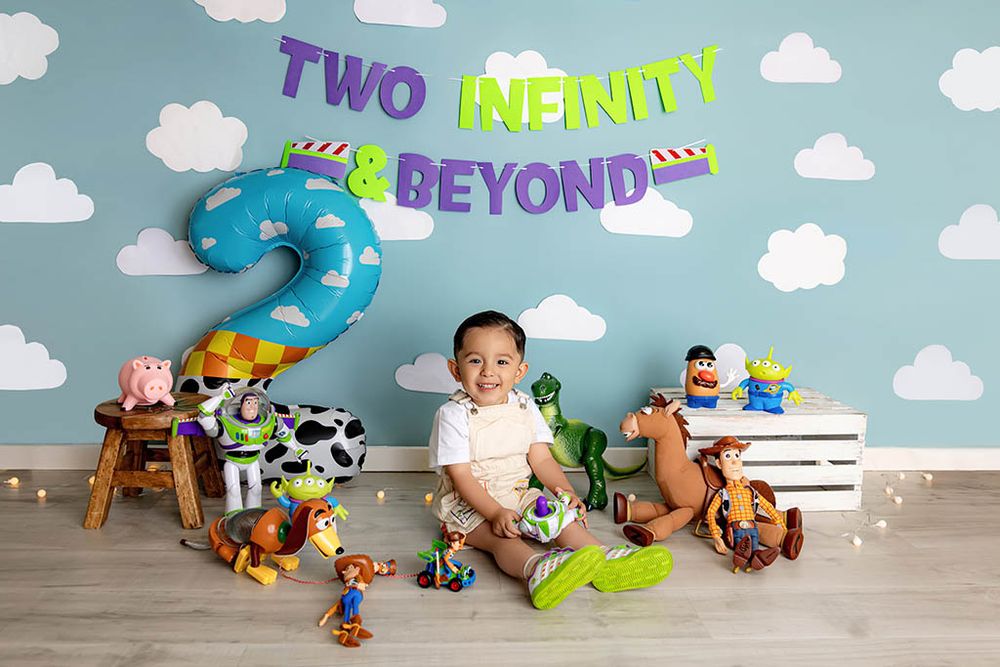 Toddler boy sits in front of Toy Story themed second birthday backdrop with balloons, toys, and Two Infinity and Beyond banner.