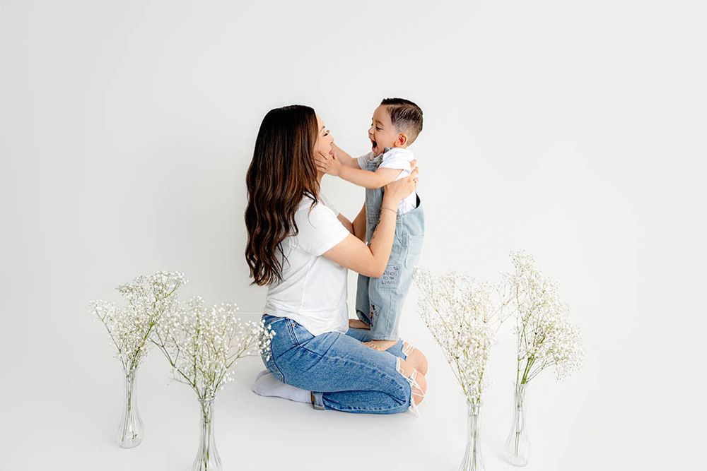 A mother lovingly lifts her smiling toddler boy in denim overalls among white floral arrangements.