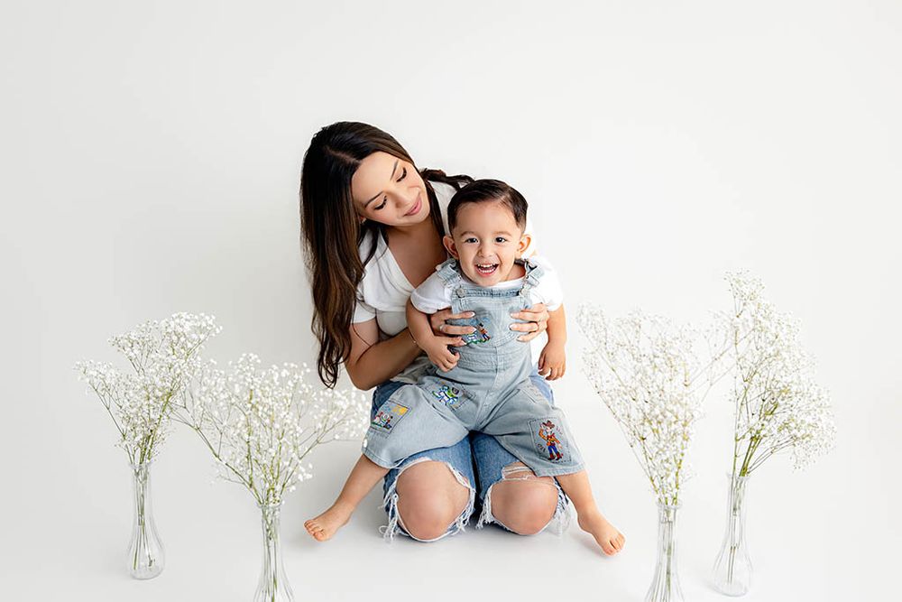 A mother hugs her smiling toddler boy in denim overalls among white flowers on a white background.