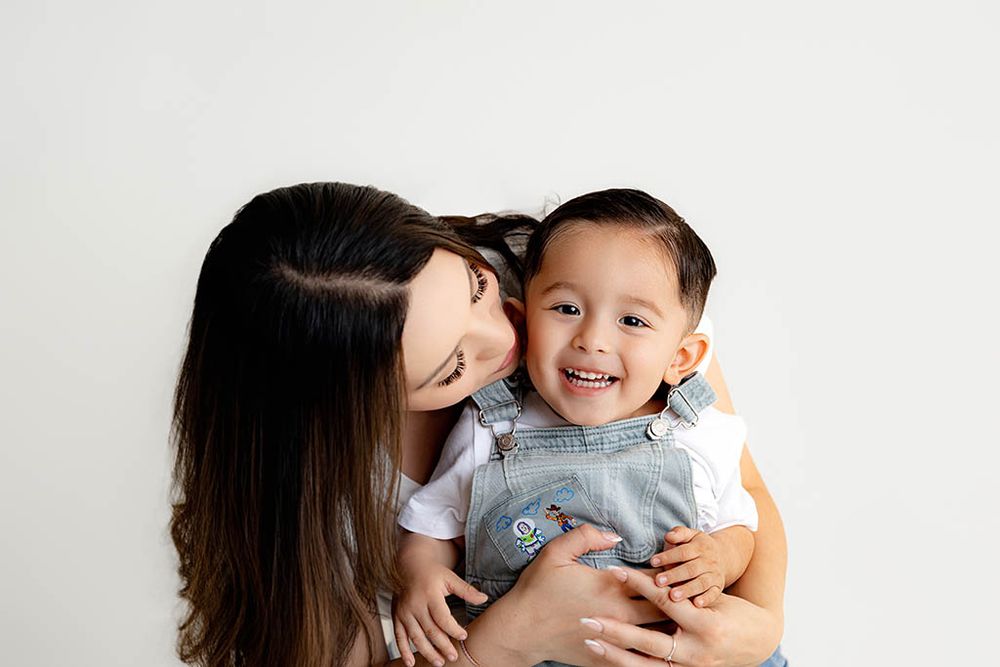 A loving mother kisses her giggling toddler son wearing denim overalls in a studio.