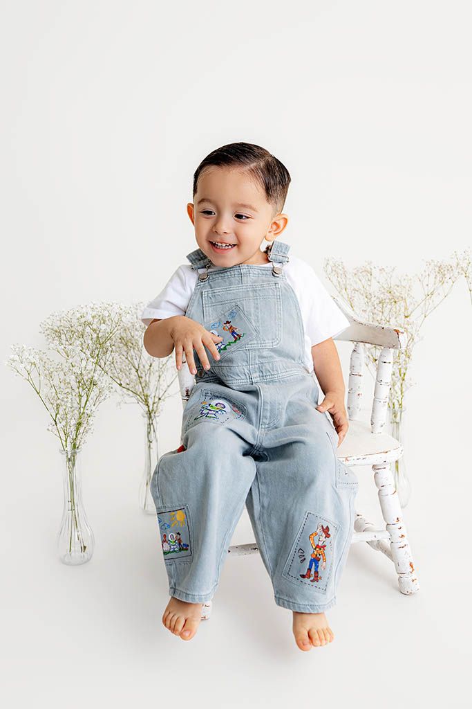 A happy toddler boy in Toy Story denim overalls sits on a white chair surrounded by white flowers.