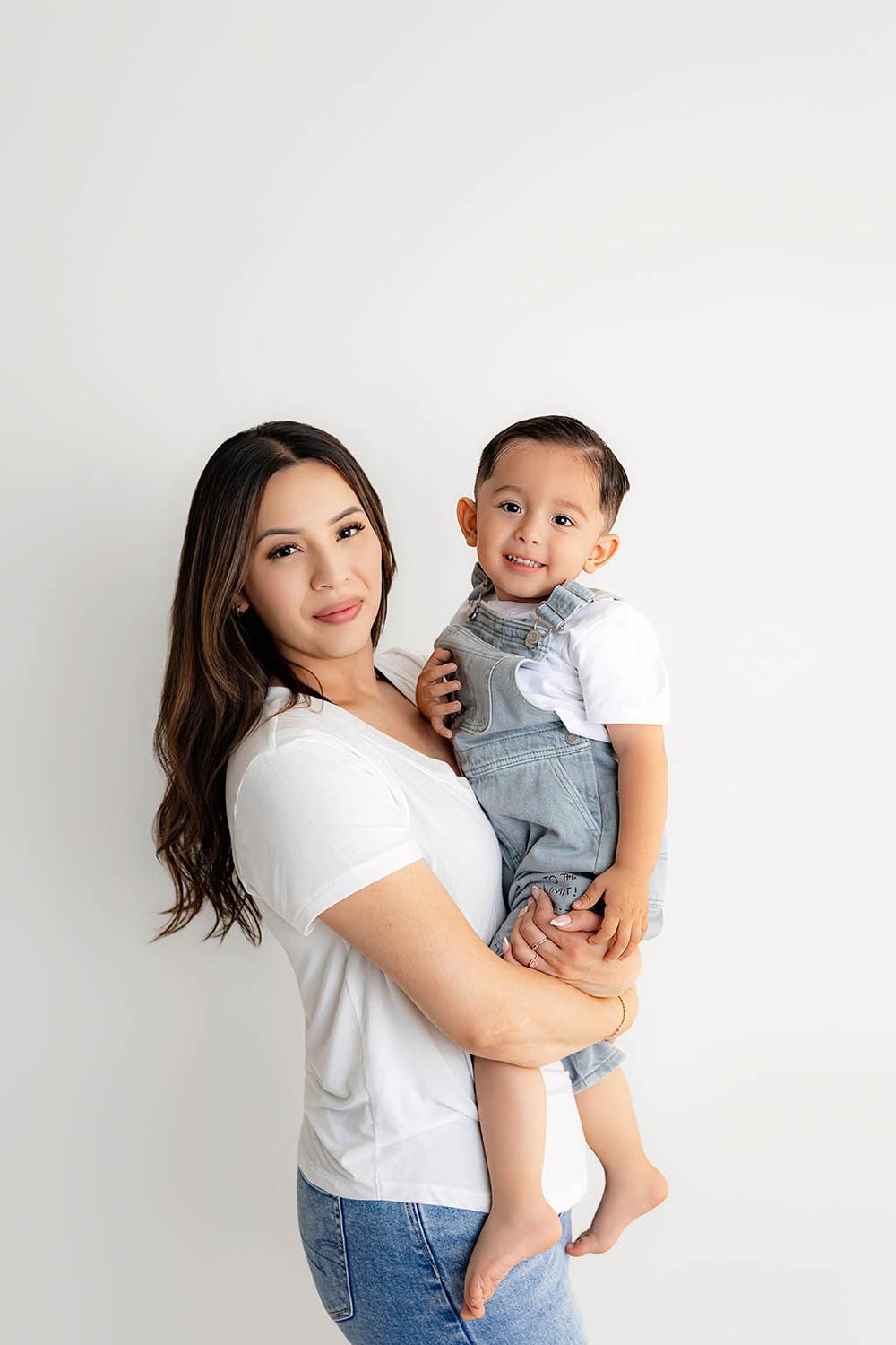 A young Asian mother in white tee and jeans holds her smiling toddler boy in denim overalls against white background.