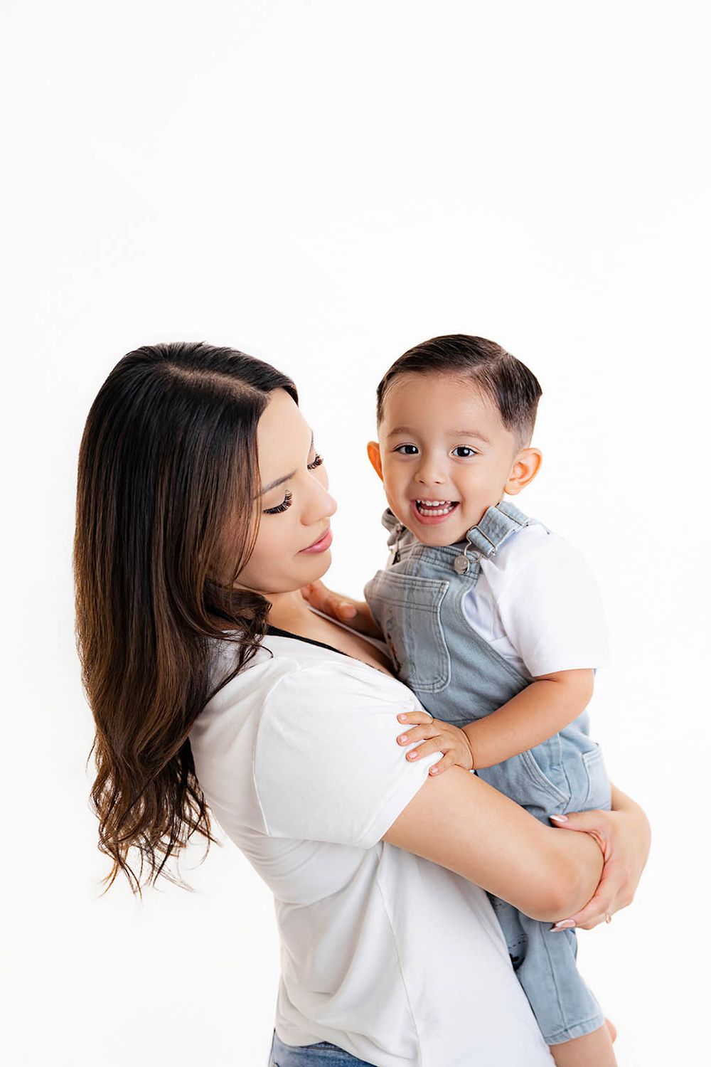 A mother holds her smiling toddler boy in denim overalls against a white background.