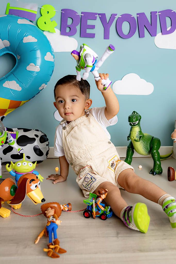 Toddler boy in overalls plays with Buzz Lightyear toy at Toy Story themed birthday party with characters and decorations.
