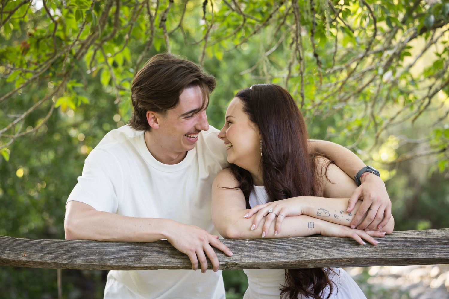 A young couple smiles at each other while leaning on a wooden fence surrounded by lush green trees.