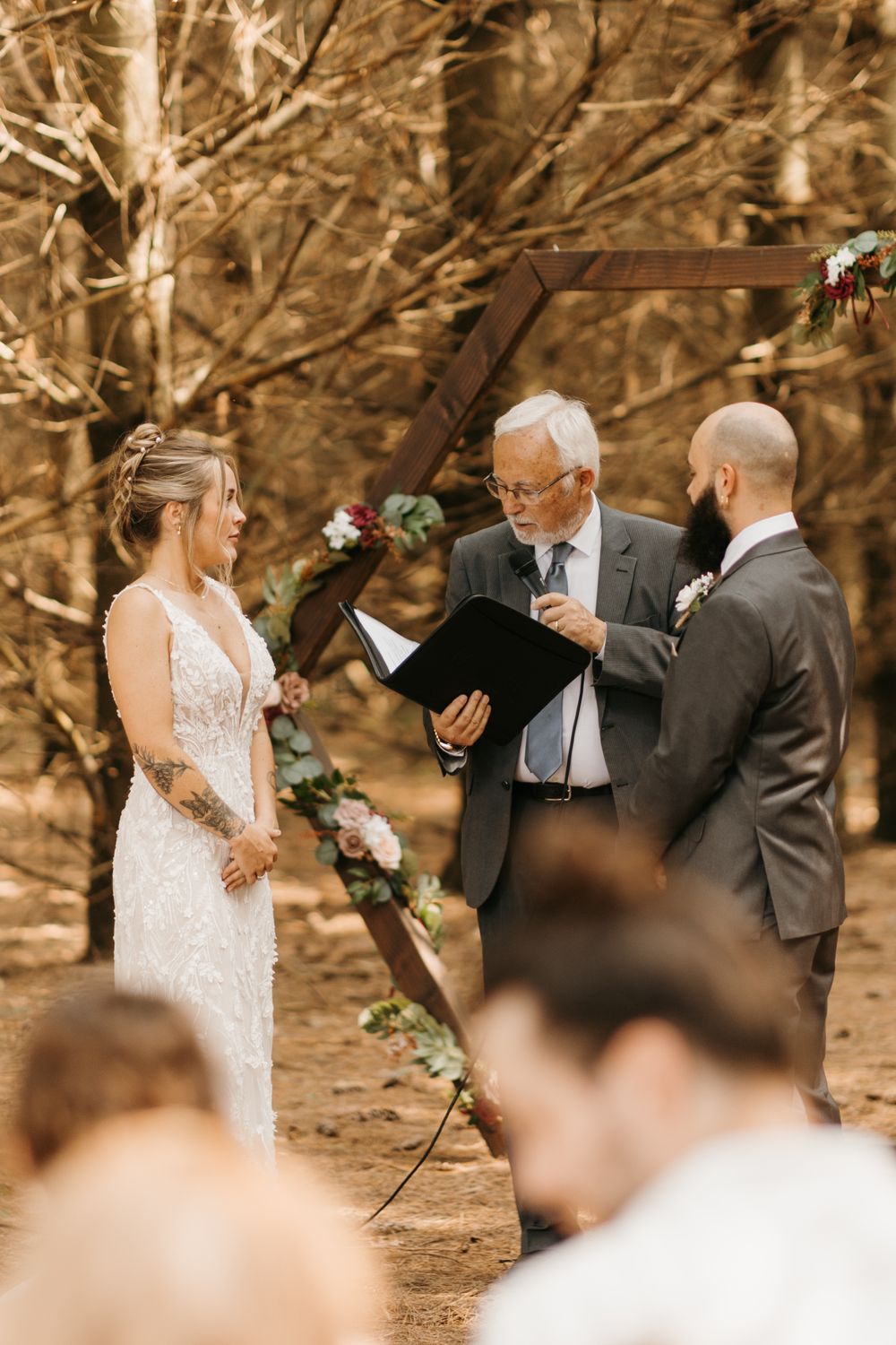 Outdoor woodland wedding ceremony with bride, groom, and officiant under a hexagonal wooden arch adorned with floral greenery.