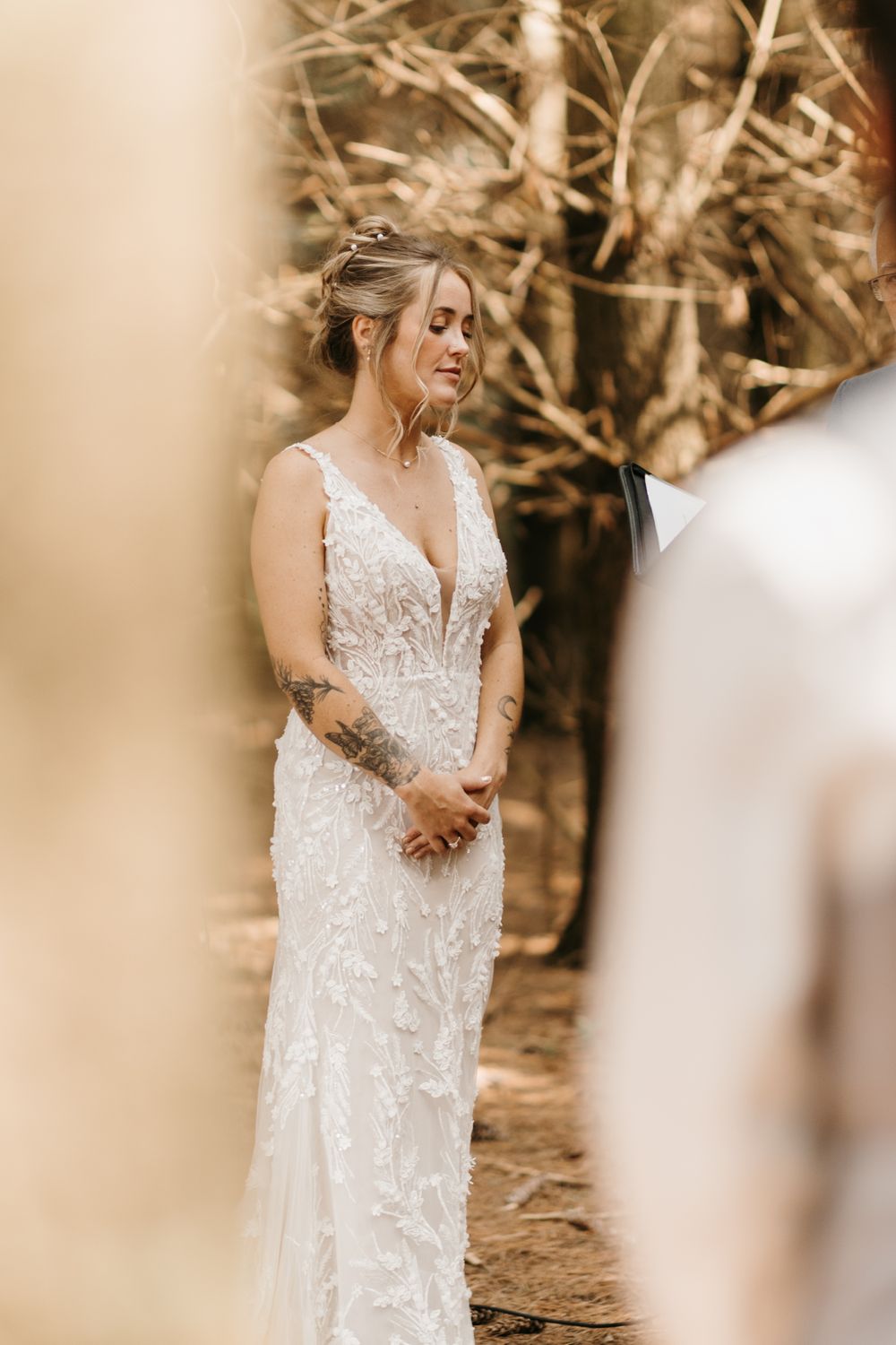 Emotional bride in a lace floral wedding gown with tattoos, hands clasped during an outdoor forest wedding ceremony.