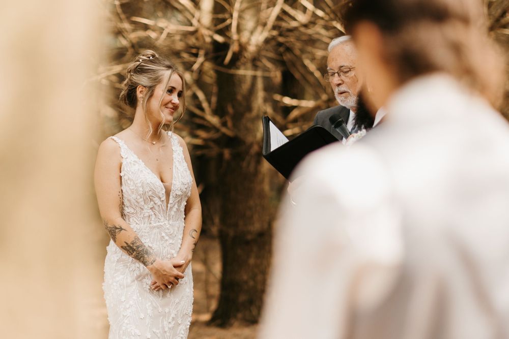 Smiling bride in a white lace gown listens to the officiant during a romantic outdoor woodland wedding ceremony.