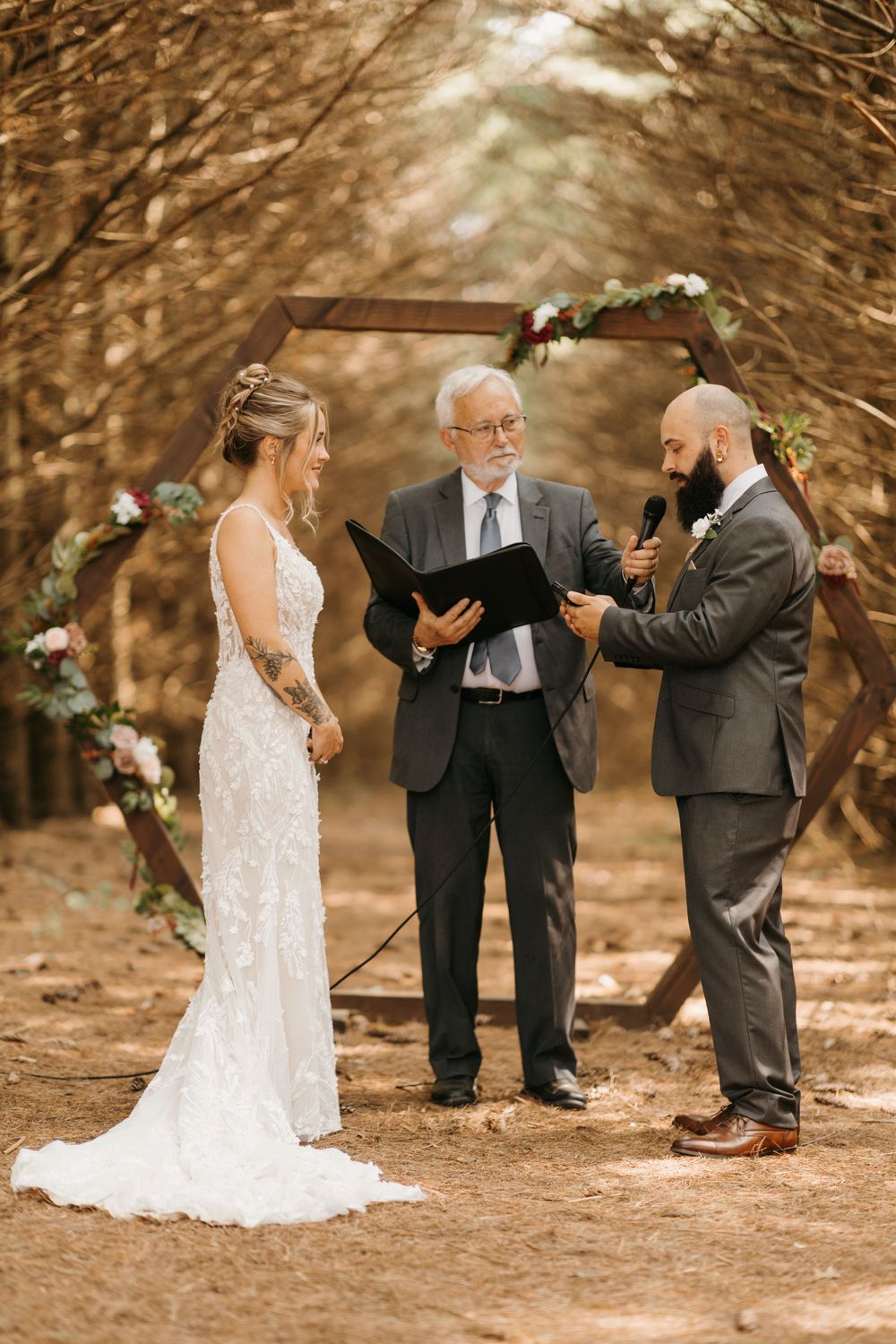Groom reads wedding vows holding a microphone beside his bride and officiant under a rustic hexagonal wooden arch.