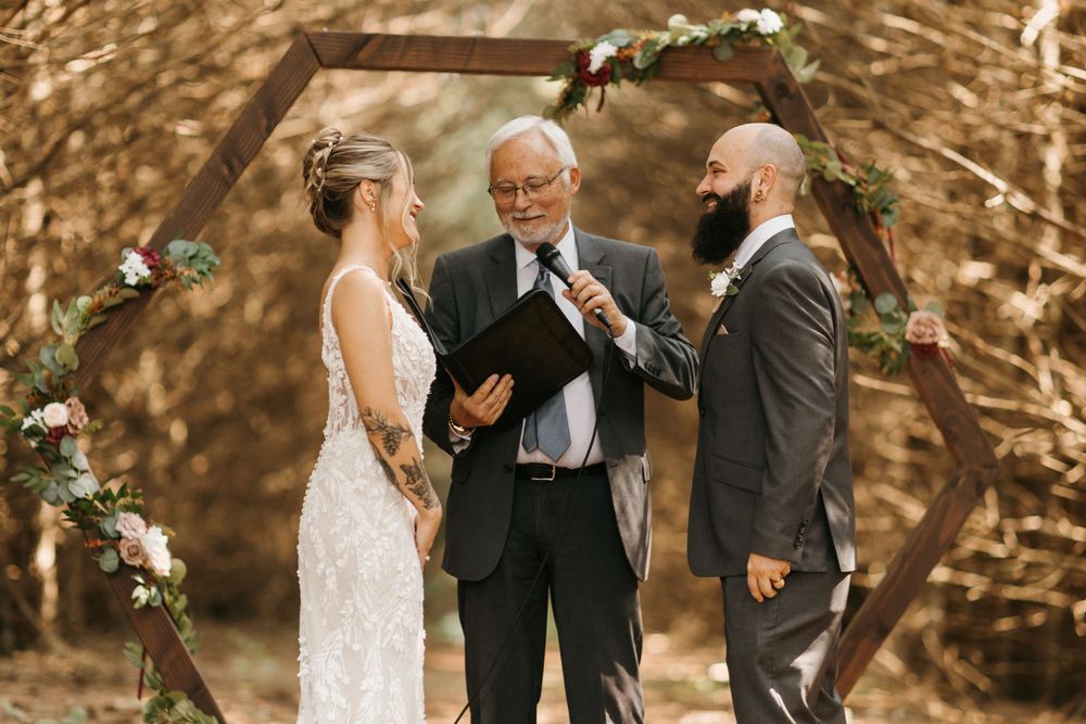 Bride and tattooed groom smile during outdoor wedding ceremony beneath a wooden hexagonal arch with floral decorations.