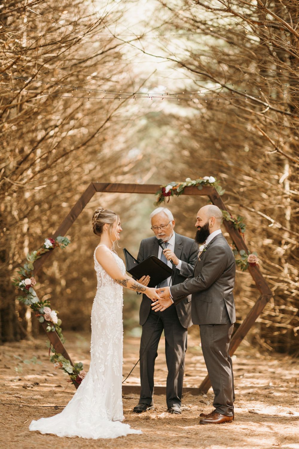 Couple exchanges rings with officiant reading from a black book during a forest wedding under a wooden arch.