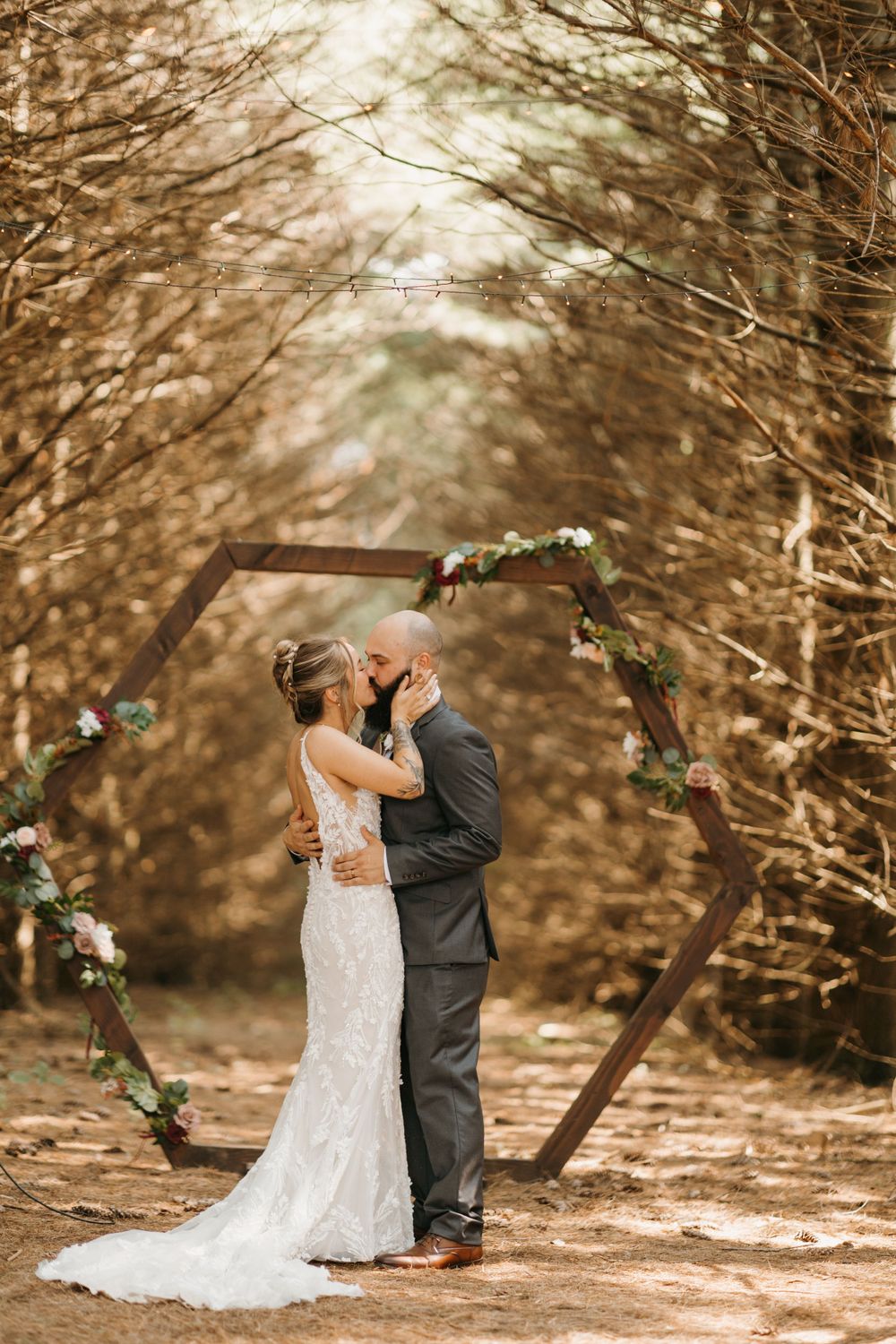 Newlyweds share their first kiss under a rustic hexagonal wooden arch surrounded by autumn trees in a forest.