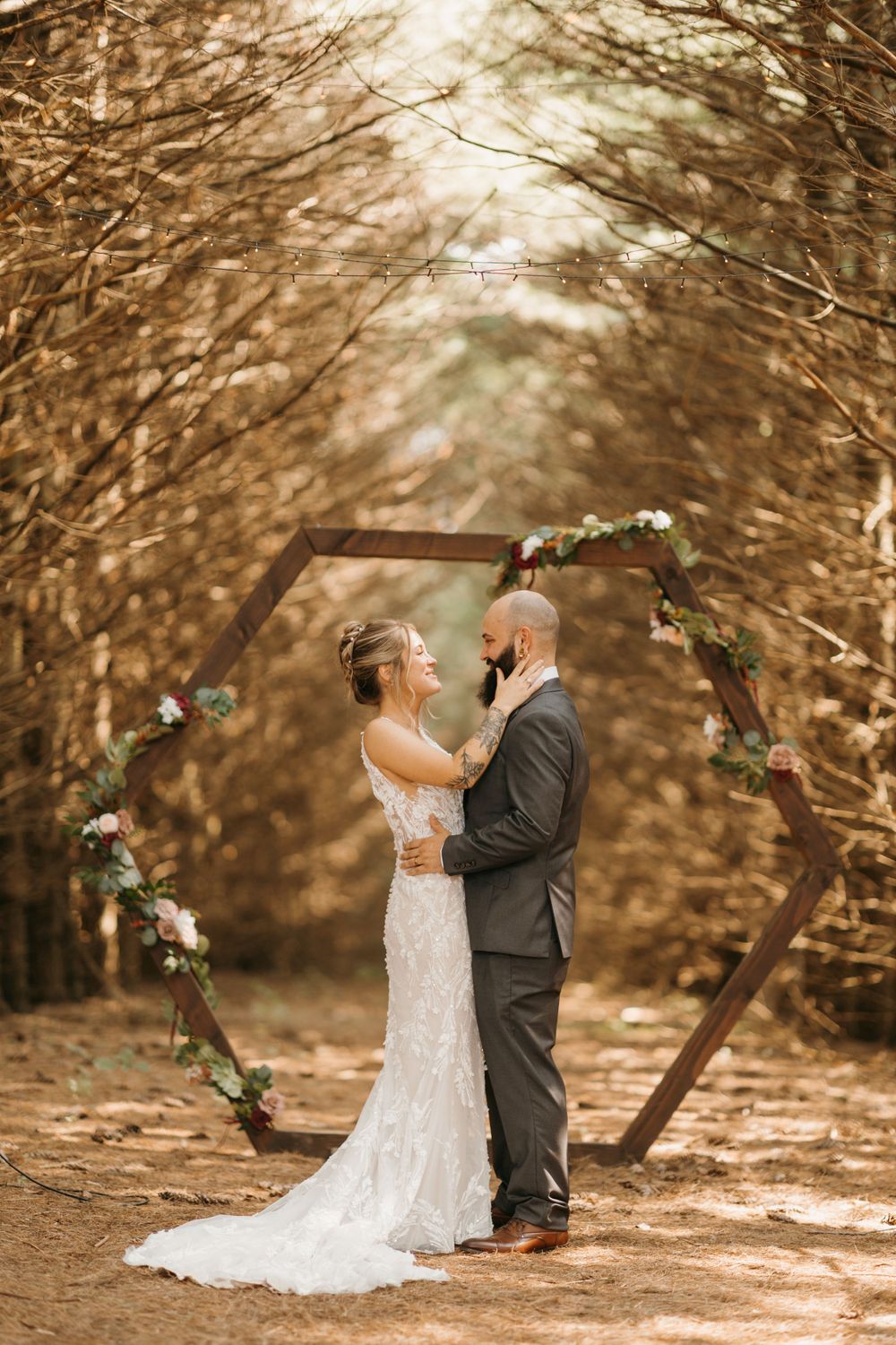 Bride and groom embrace romantically under a hexagonal wooden wedding arch decorated with greenery in an autumn forest.