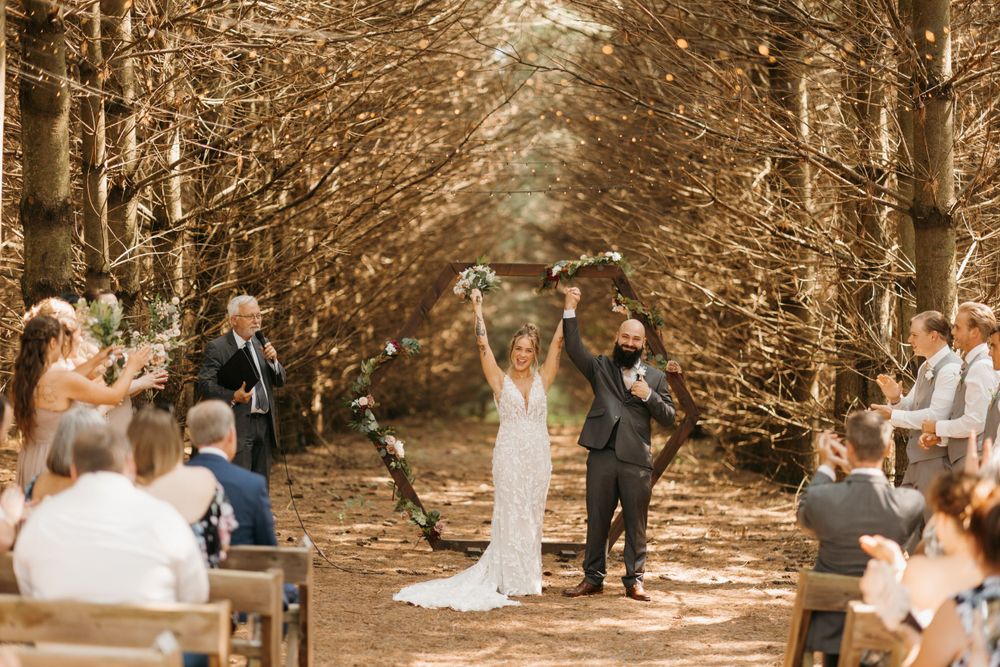 Newlyweds celebrate joyfully raising their arms as they walk back down the aisle in a tree-lined outdoor venue.
