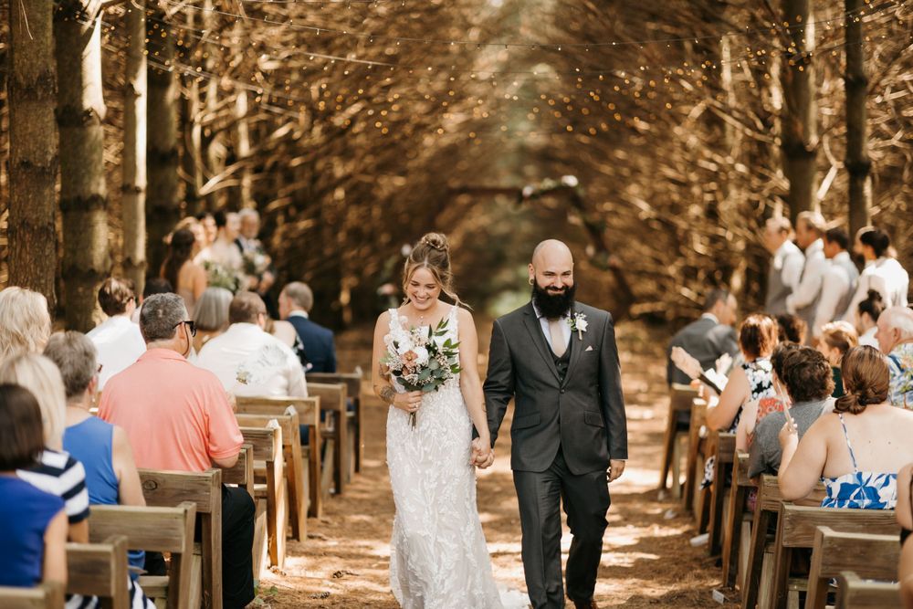 Bride and groom walk hand-in-hand down a tree-lined outdoor wedding aisle as guests look on.