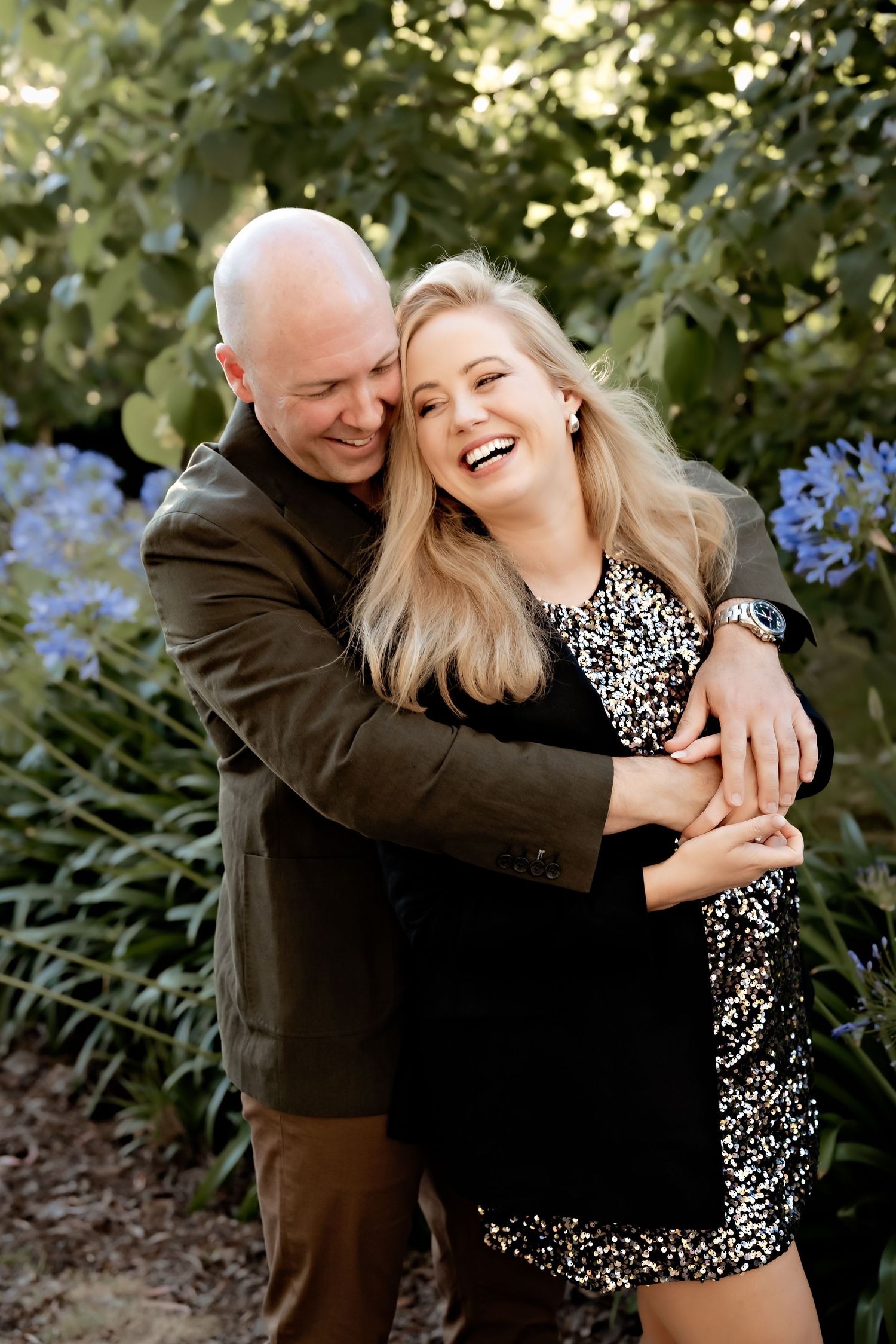 A couple shares a joyful embrace in a garden with blue flowers in the background.