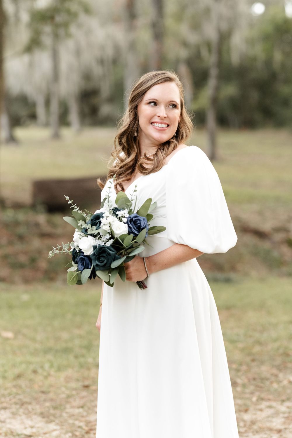 A joyful bride in a white winter wedding dress holds a beautiful bouquet with navy and white flowers outdoors.
