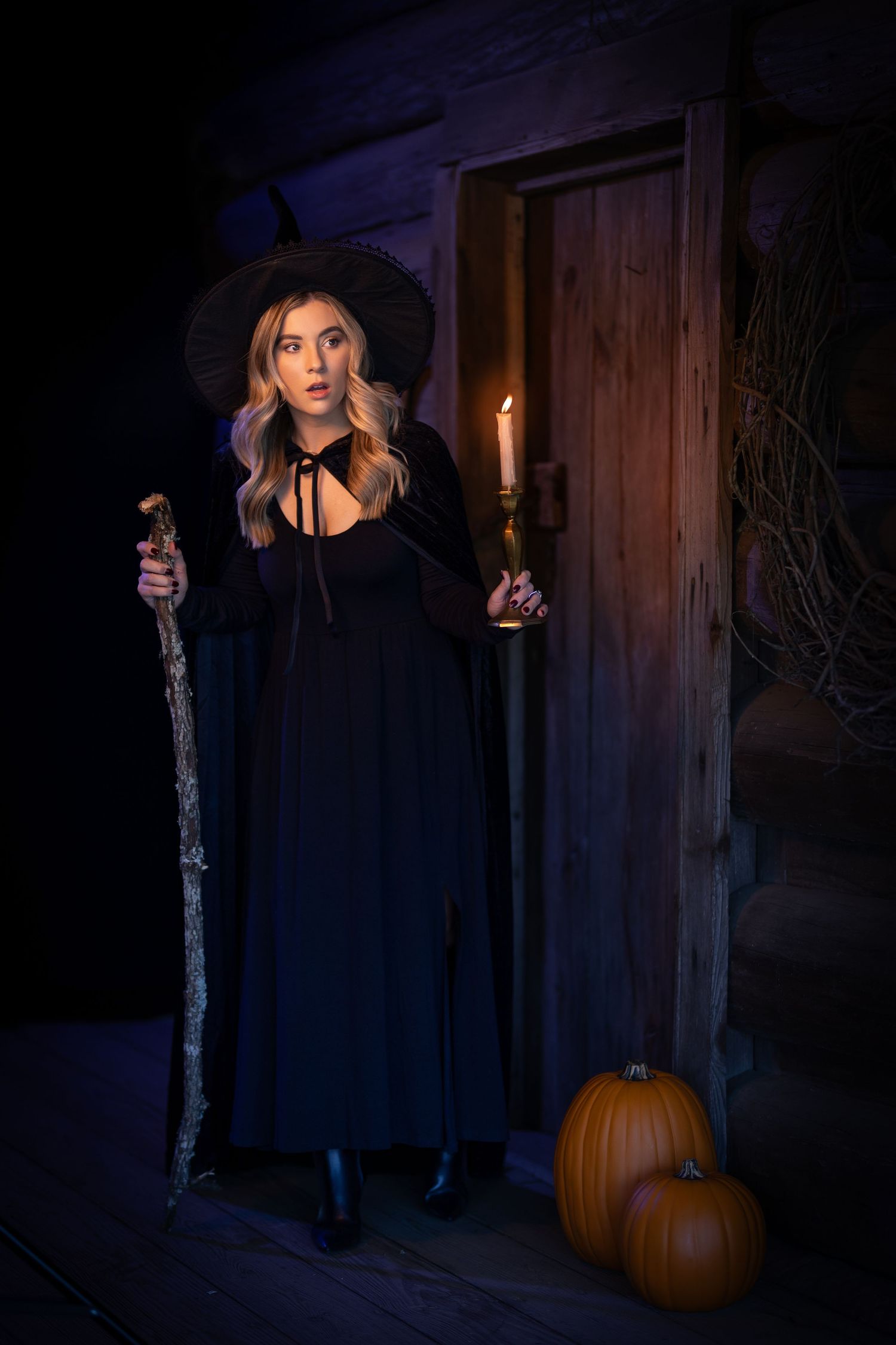 A witch in a black dress and hat stands in a doorway at night holding a staff and candle, with pumpkins by her feet.