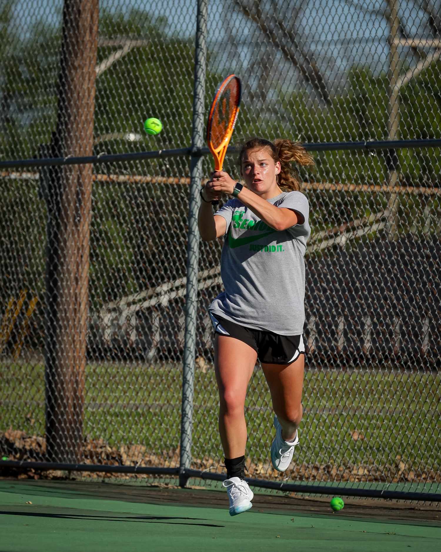 Tennis player in gray shirt hits backhand shot during practice on outdoor court with green balls scattered nearby.