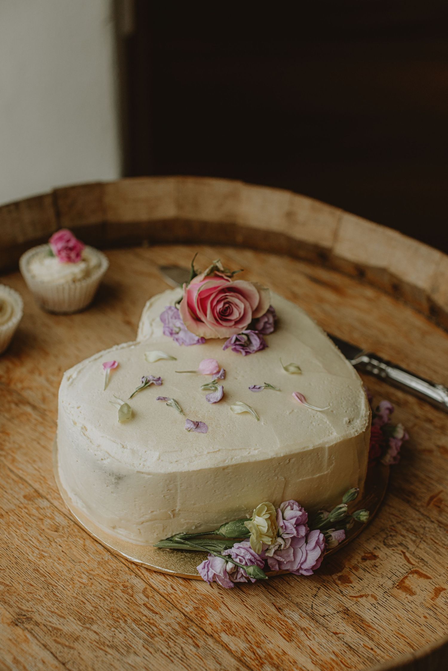 Heart-shaped wedding cake decorated with delicate pink and purple roses on rustic wooden surface.