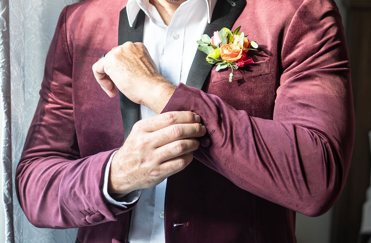 Groom in burgundy velvet blazer adjusting cufflinks with floral boutonniere on lapel.