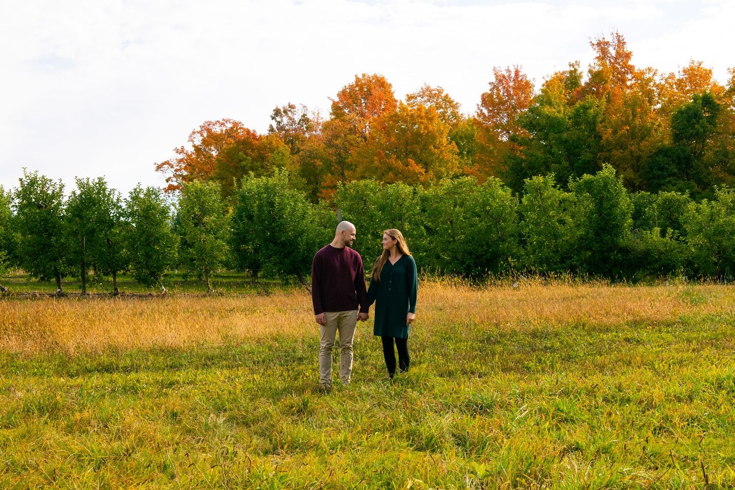 Two people stand together in a golden autumn field with colorful fall foliage in the background during engagement photos