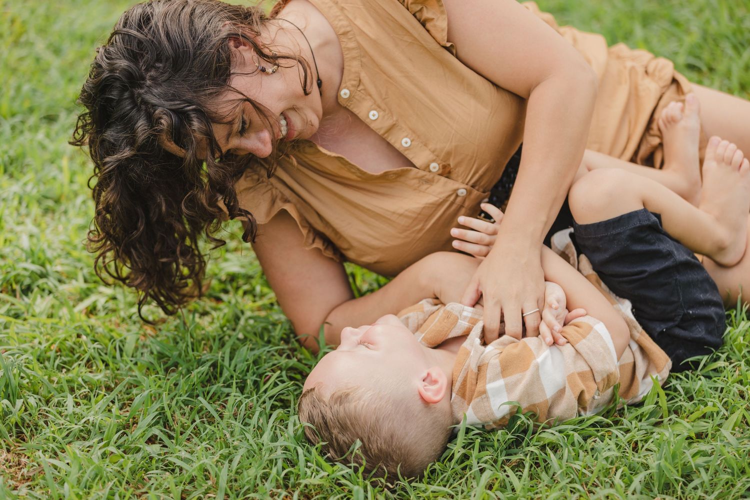 A parent and child share a tender playful moment while lying on green grass in warm sunlight.