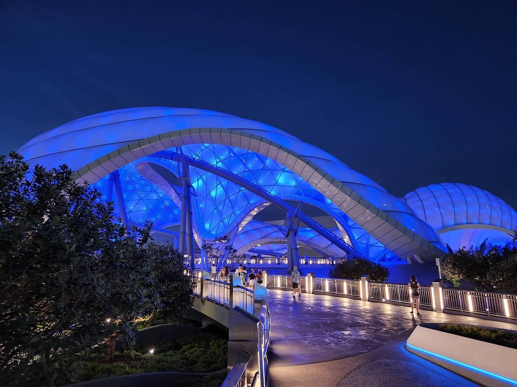 A modern architectural bridge illuminated with blue lighting at night creates a striking reflection on the water below.