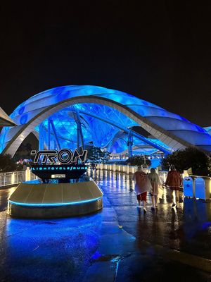 Illuminated TRON attraction at night with blue neon lights and curved architectural elements reflecting on wet pavement.