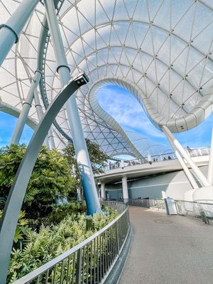 Modern architectural glass and steel canopy with curved geometric patterns frames a view of blue sky at a botanical garden entrance.