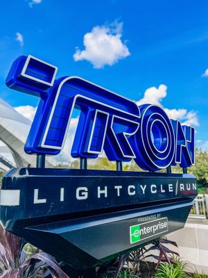Large blue TRON sign against bright blue sky showcases futuristic theme park ride entrance at Disney World.
