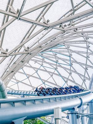 A roller coaster track viewed from below shows white architectural supports and blue rails against a bright sky.