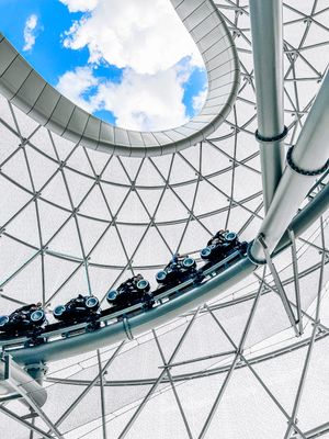 Roller coaster track curves beneath a modern geometric glass dome with blue sky visible through the transparent roof.