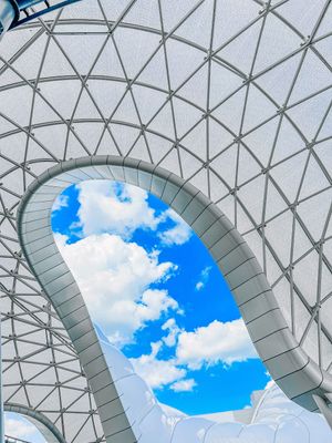 Geometric glass dome ceiling with triangular patterns frames a view of bright blue sky and white puffy clouds.