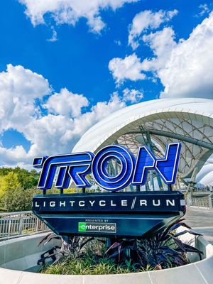 TRON Lightcycle Run glowing blue sign and white coaster track against bright blue sky at Disney World theme park.