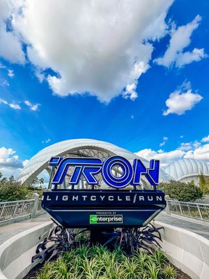 The TRON Lightcycle Run attraction sign against a bright blue sky with white clouds at Disney World theme park.