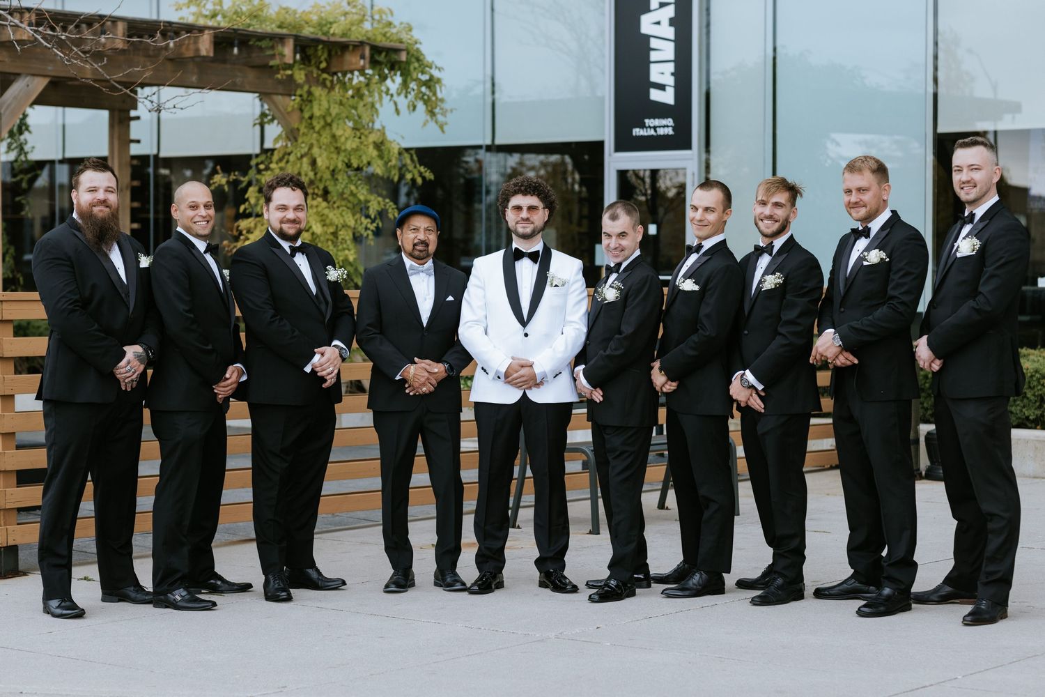 Wedding party in black tuxedos and one white tuxedo jacket pose together outside a modern building.