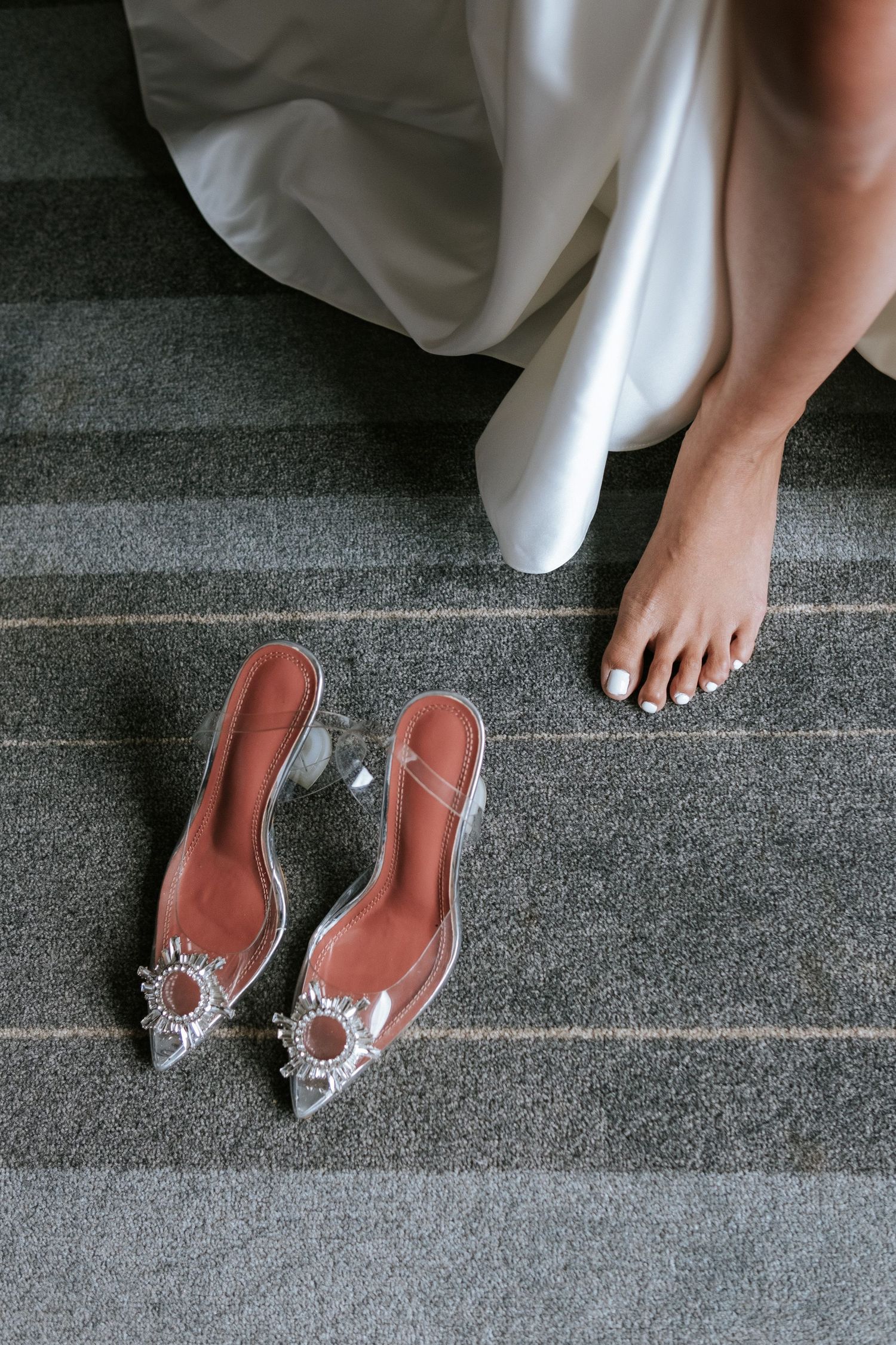 A wedding dress hem and barefoot extend down grey carpeted stairs beside elegant crystal-embellished shoes.
