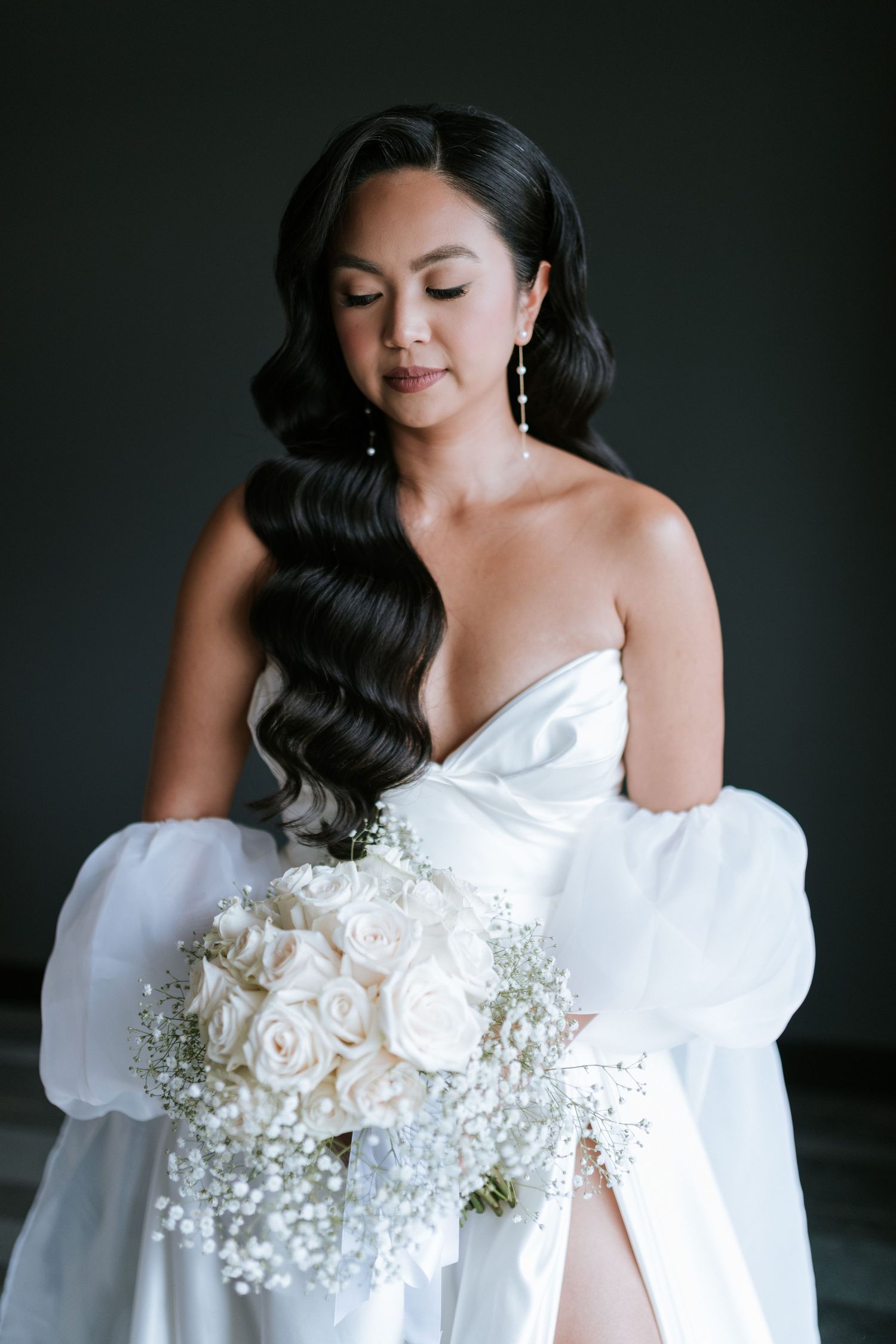 A bride holds a beautiful white rose and baby's breath bouquet while wearing an elegant strapless gown.