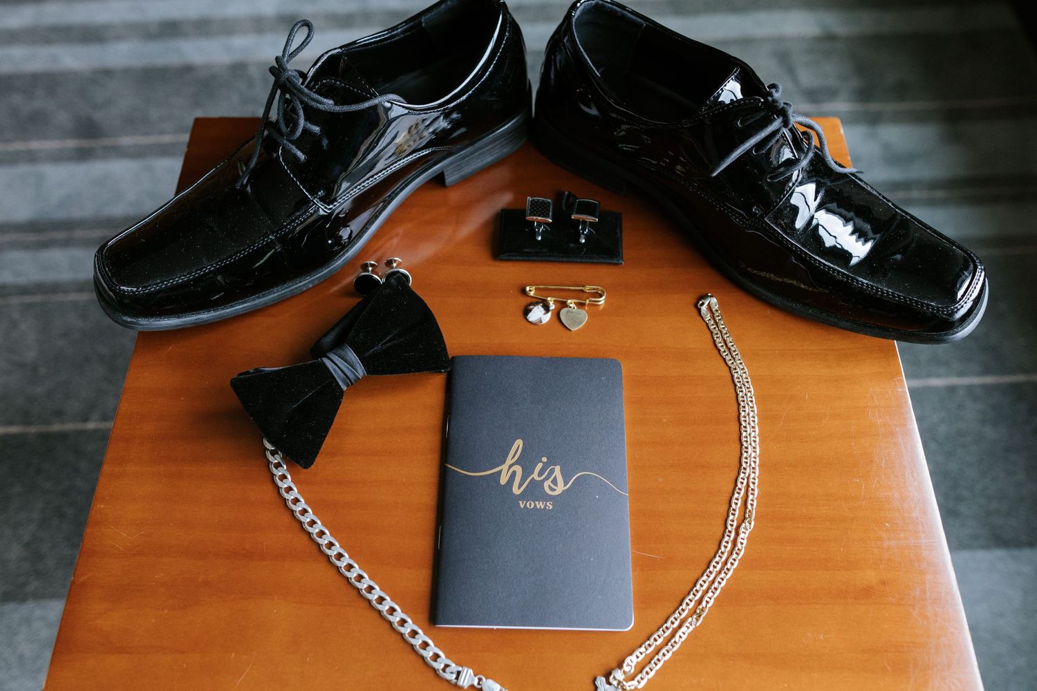 Wedding accessories including black dress shoes, bow tie, cufflinks and jewelry arranged on wooden surface.