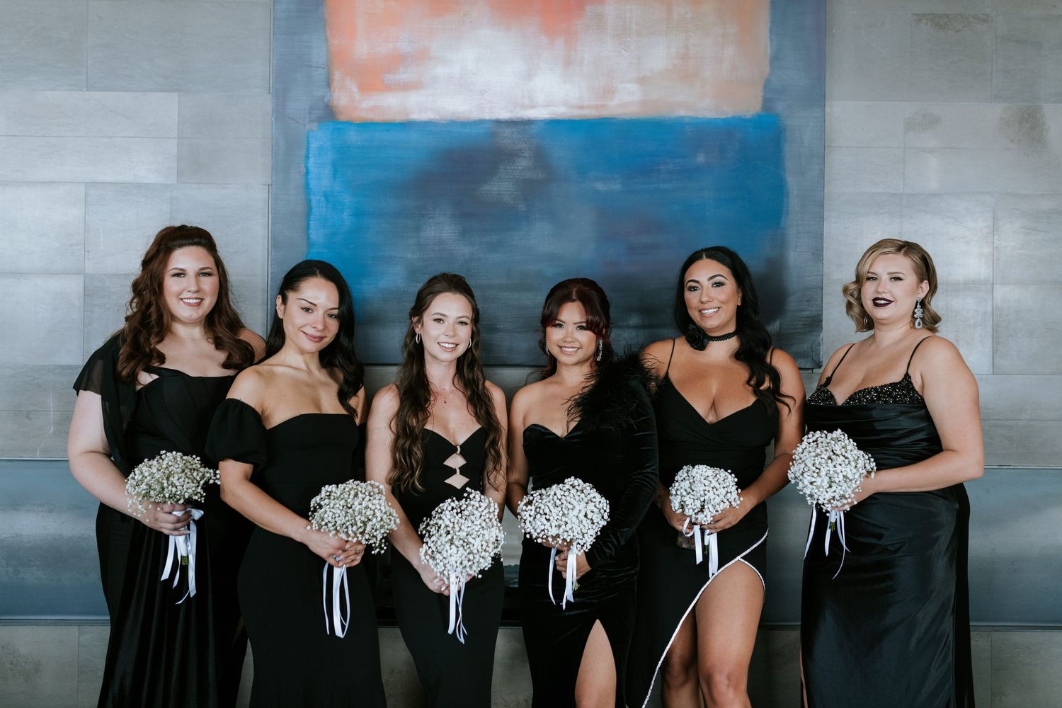 Bridesmaids in elegant black dresses hold matching white bouquets while posing against a blue and coral backdrop.