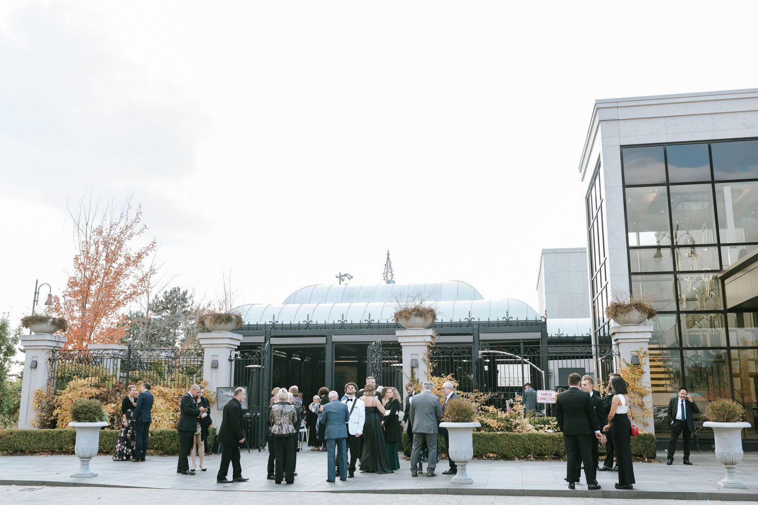 Wedding guests mingle outside a modern glass building with autumn trees in the background.