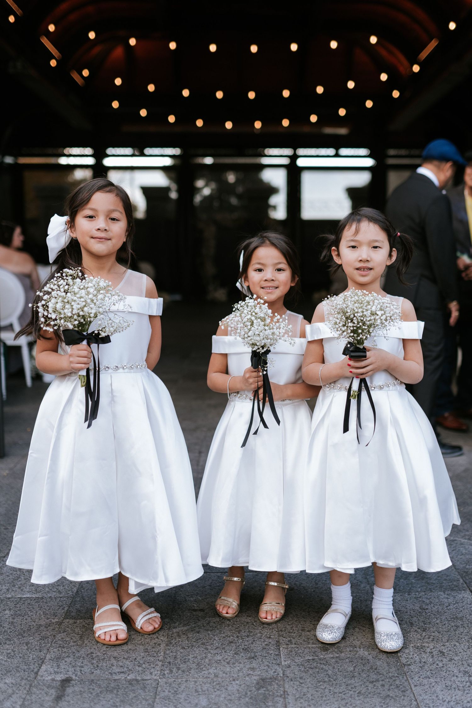Three flower girls in matching white dresses hold baby's breath bouquets under string lights.