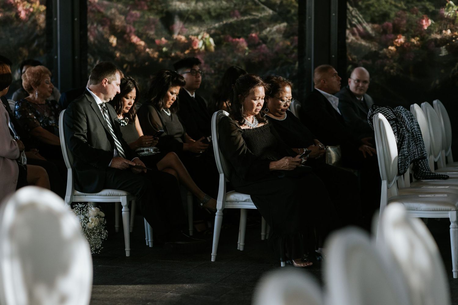 Wedding ceremony guests seated in white chairs under dim lighting during an indoor service.