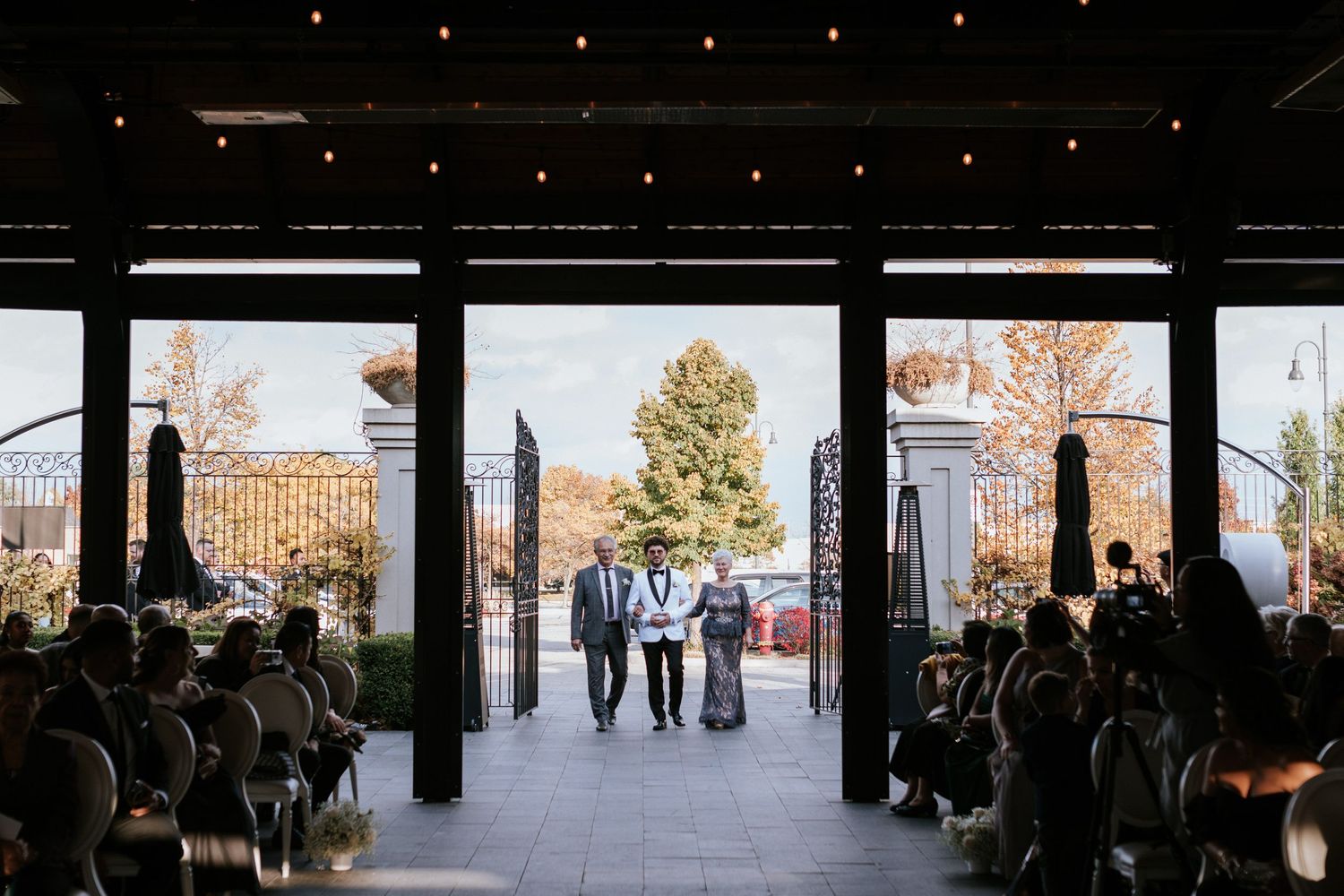 Wedding guests gather in a modern venue with large windows and string lights overhead.