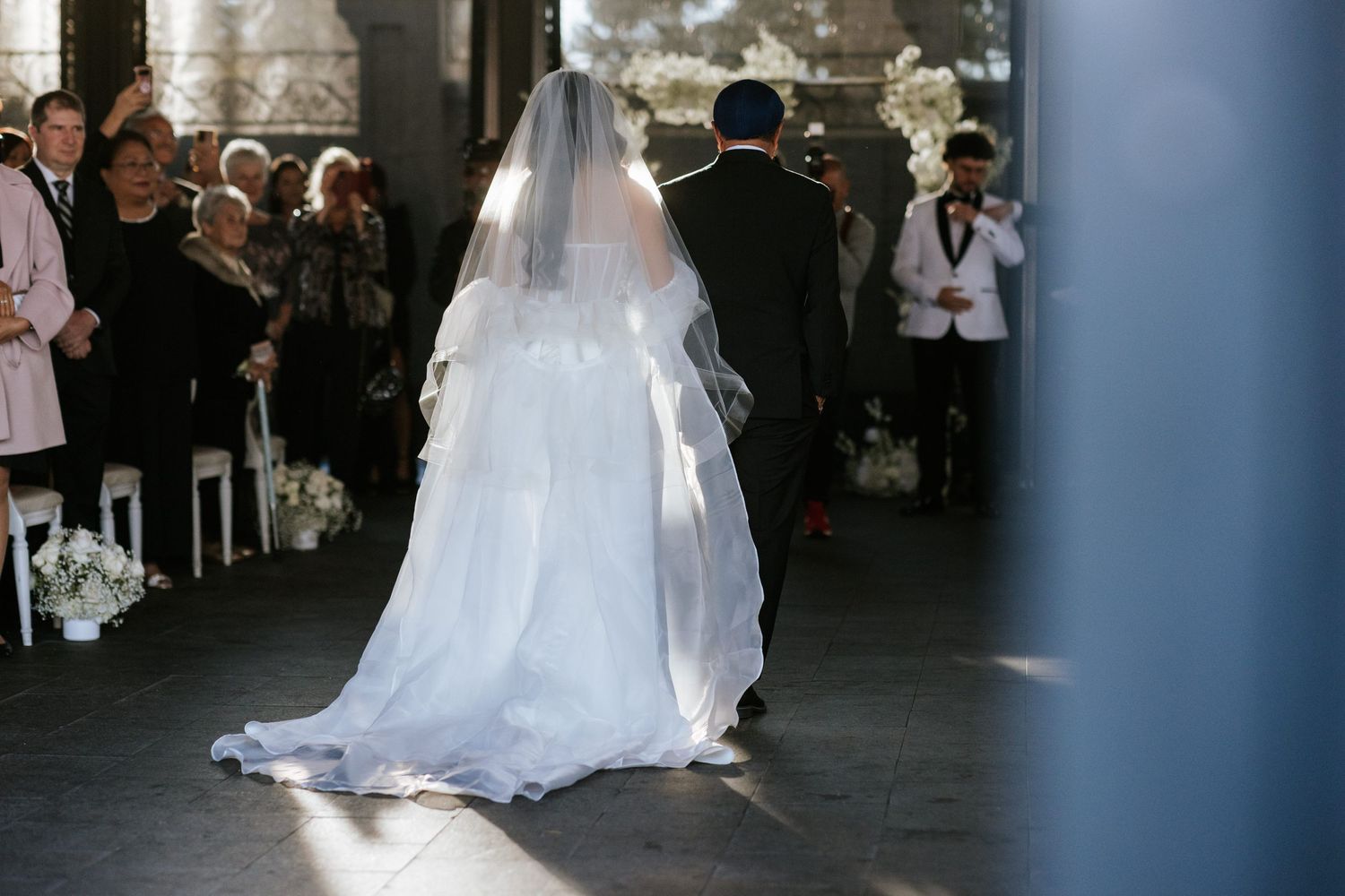A bride in a flowing white wedding gown and veil stands at the ceremony venue with guests looking on.