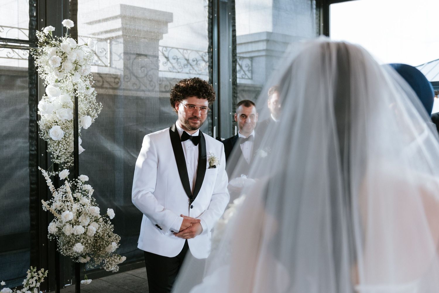 A grand wedding ceremony setup with elegant white floral arrangements and a groom in a white tuxedo.