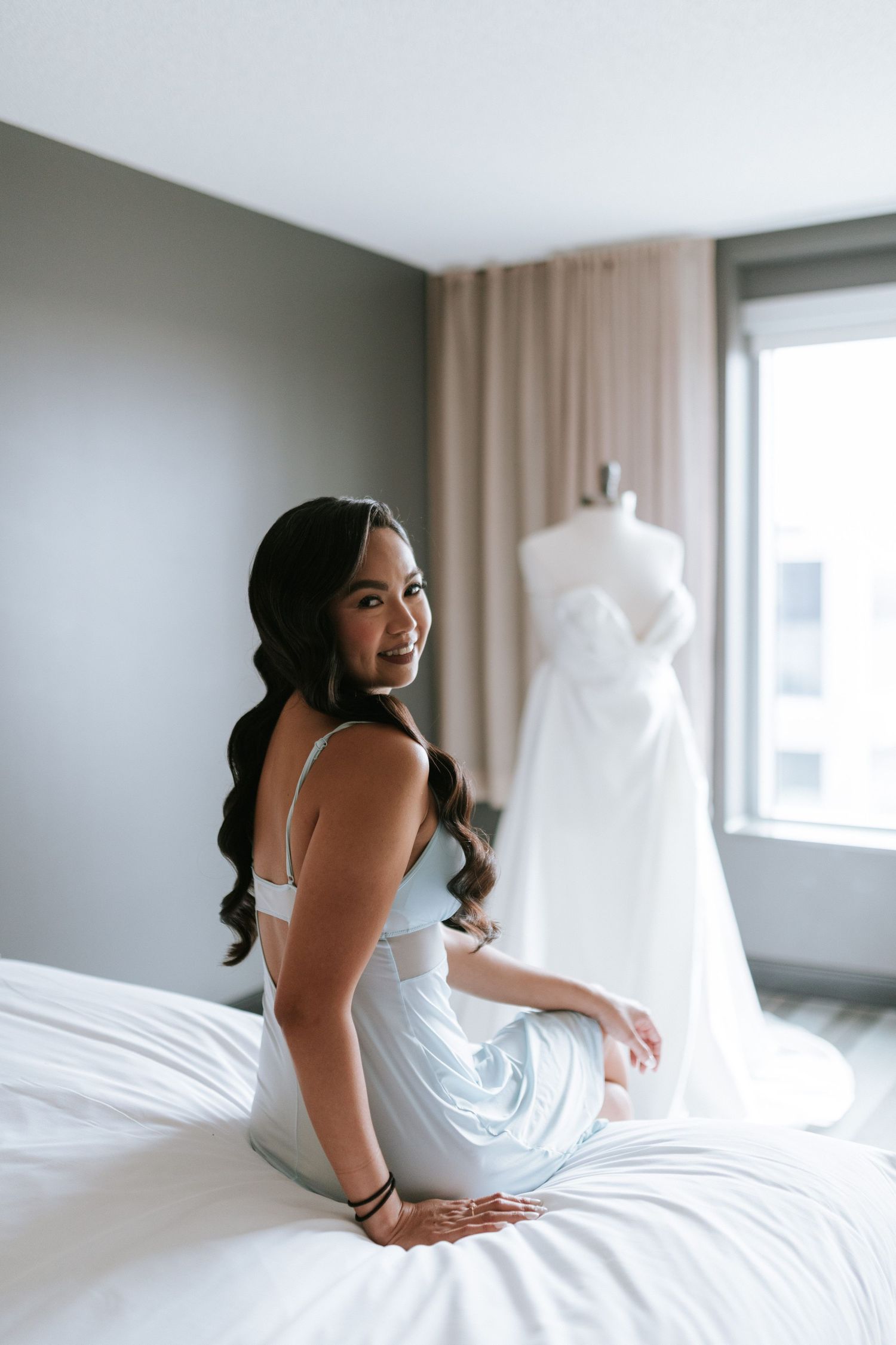 Bridal preparation scene with white dress hanging in background of modern hotel room.